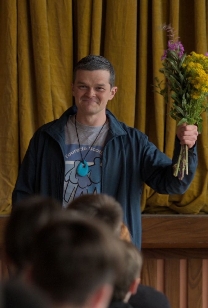 A man stands on a stage in front of an audience, holding a colourful bouquet of flowers up in one hand. They wear a dark jacket, a t-shirt with a design, and a necklace with a large pendant. The background features closed golden curtains.