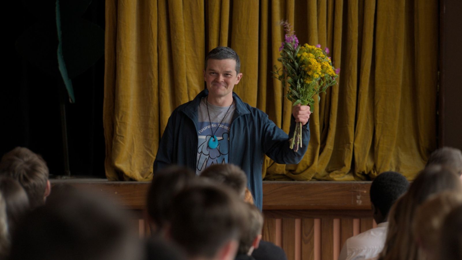 A man stands on a stage in front of an audience, holding a colourful bouquet of flowers up in one hand. They wear a dark jacket, a t-shirt with a design, and a necklace with a large pendant. The background features closed golden curtains.