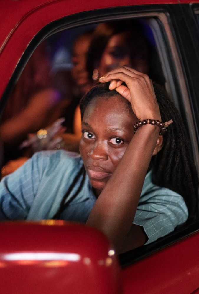 A person with long hair pulled back leans on a red car window frame, looking directly at the camera. They wear a blue checkered shirt and a wooden bead bracelet. In the background, there are blurry figures and dim, colorful lights suggesting a lively urban night scene.