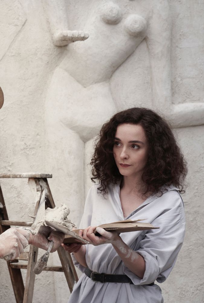 A woman with curly hair, wearing a light blouse, holds a book. She is in a workshop with plaster sculptures in the background, and a wooden ladder is visible between them.