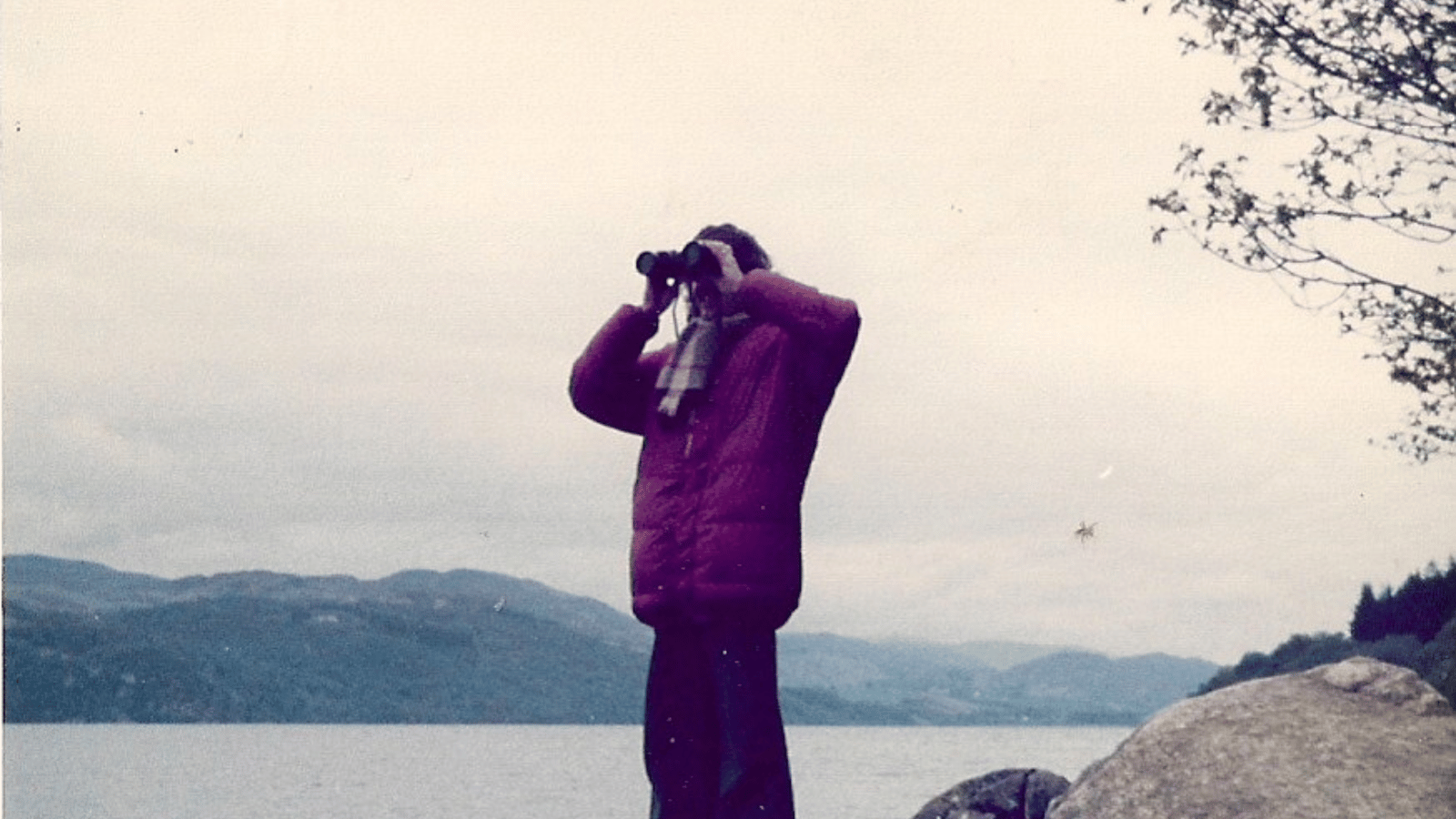 Still from Loch Ness. Person holding binoculars to their face looking out at Loch Ness,
