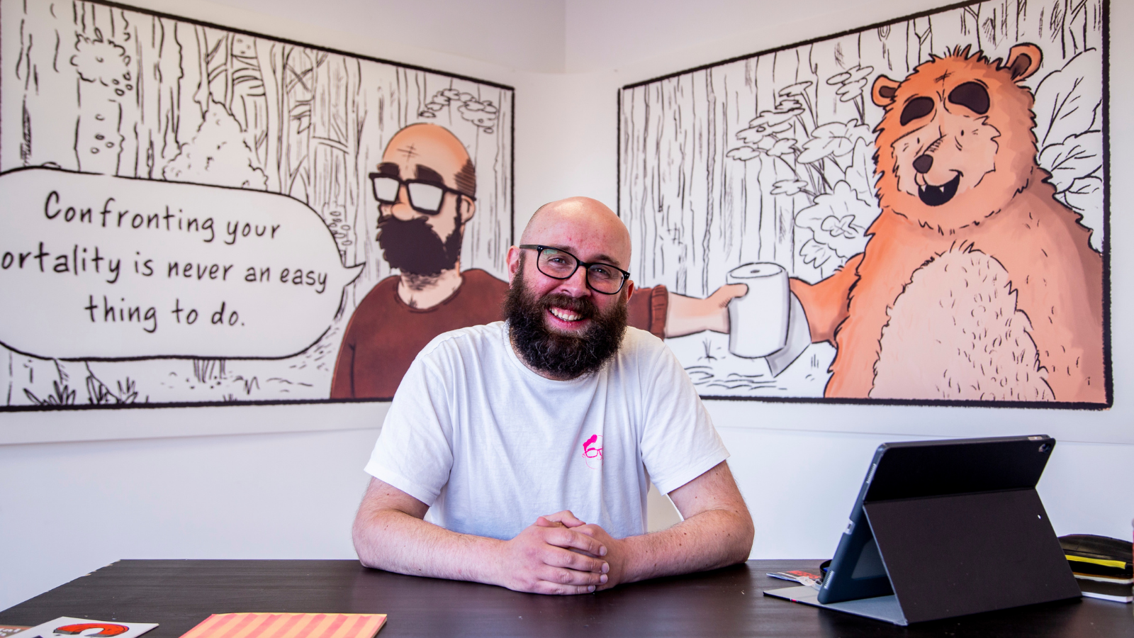 Still from Long Live My Happy Head, man sitting at a desk with comic strips on the walls behind him