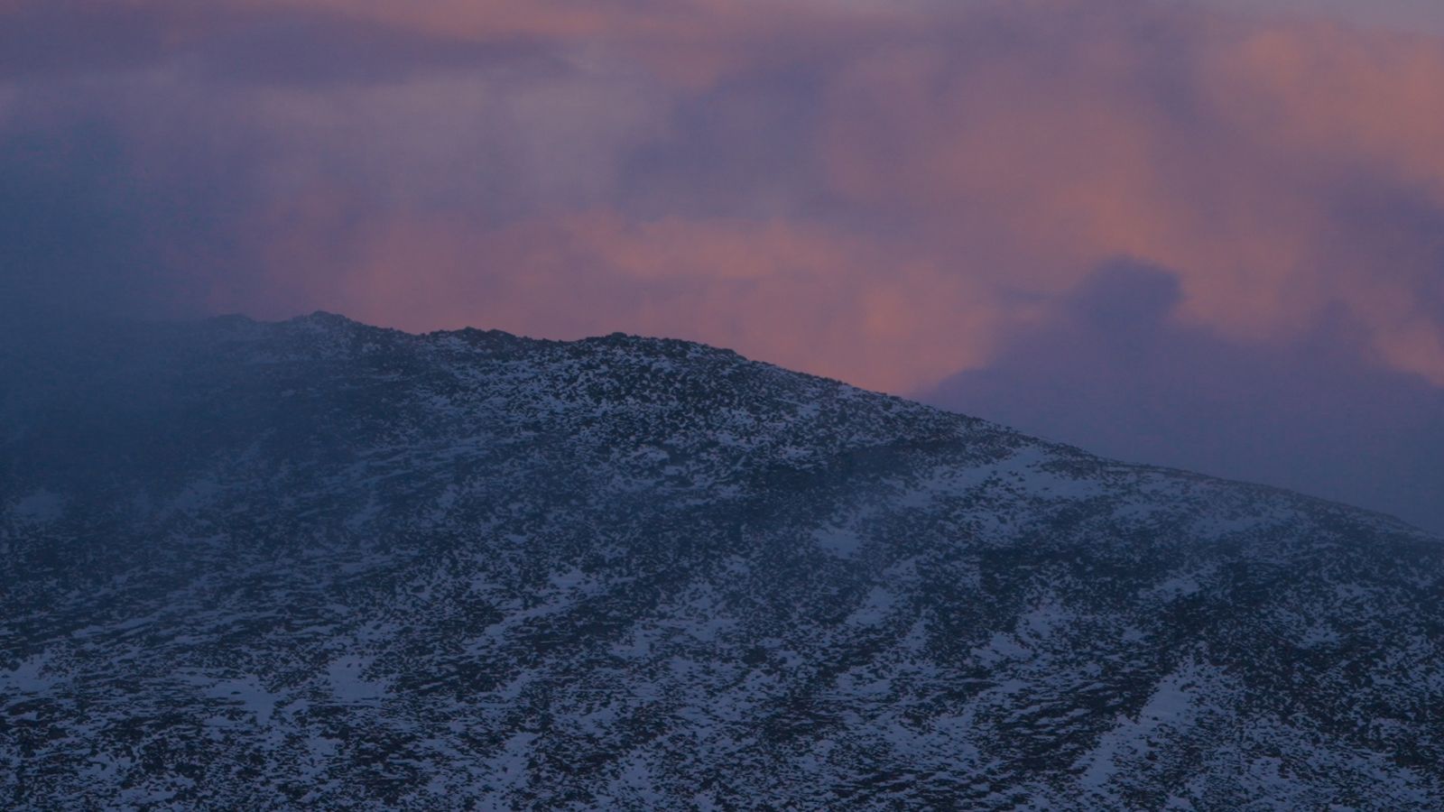 Still from Lost For Words showing a mountain in the distance with clouds overhead. The sky is a purple pink hue above.