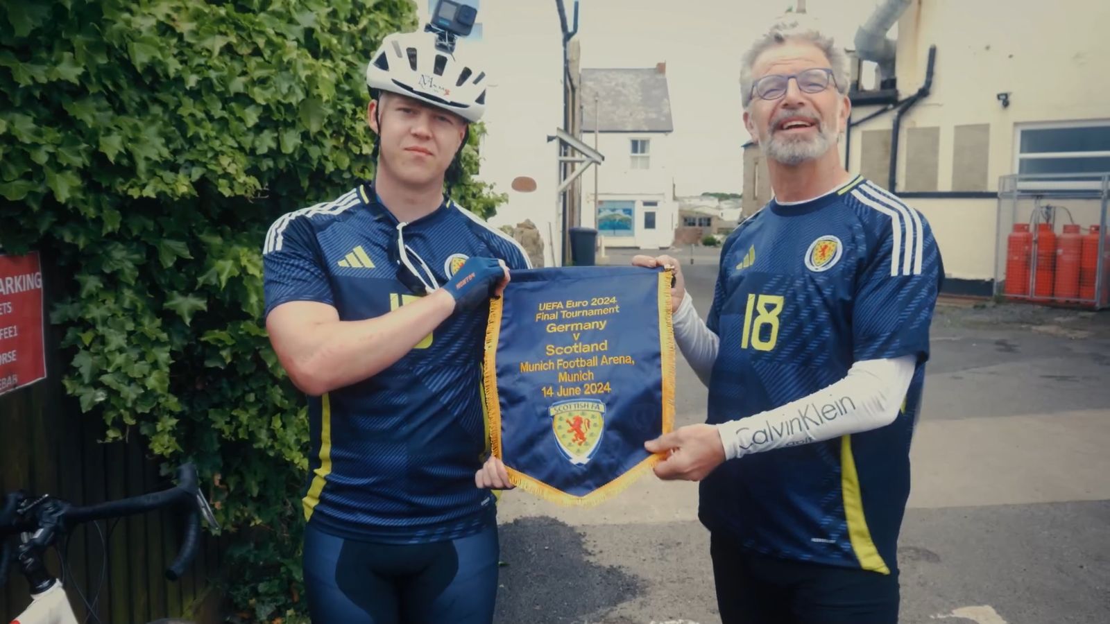 wo men wearing Scotland football strips stand side by side outdoors holding a sign.