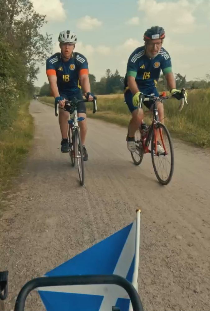 two men wearing Scotland football strips and bike helmets ride bikes side by side on a country road.