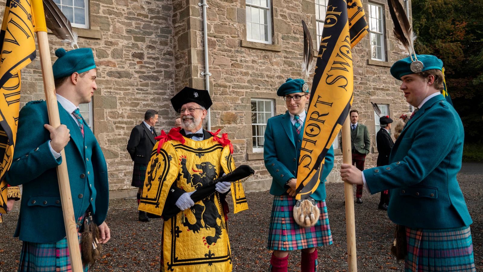 Four men stand together, three of whom wear kilts, kilt jackets and hats. Two of the men hold yellow and black banners on poles. They are standing in front of a stone house with several other people in the background.