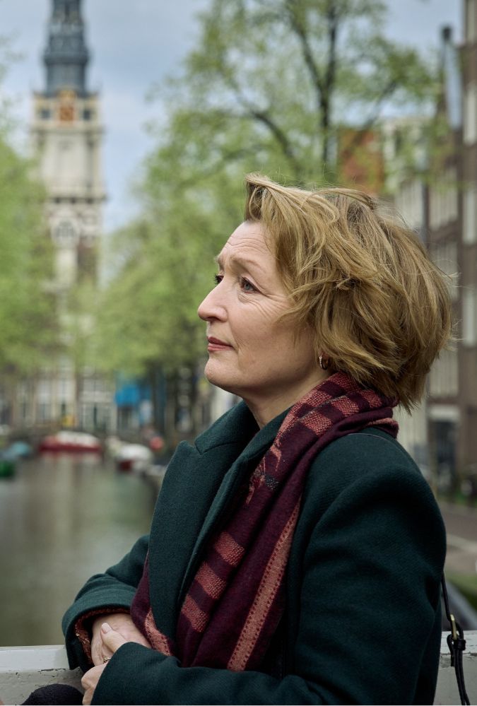 A woman stands on a canal bridge in Amsterdam, looking to the left. The background features a canal lined with green trees and a distant clock tower under a cloudy sky.
