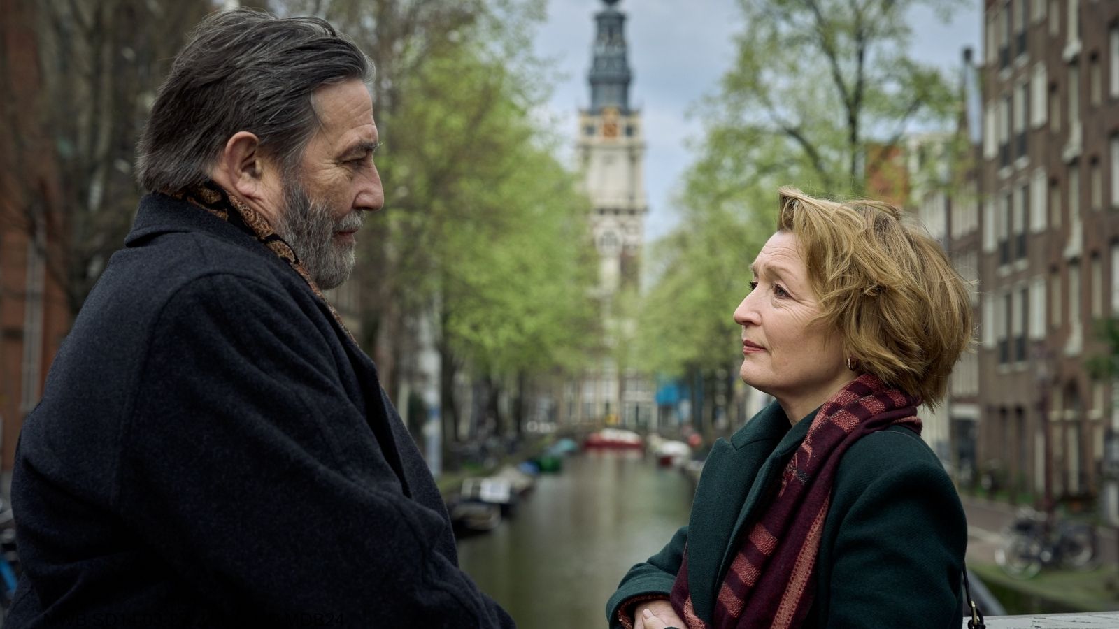 A man and a woman stand on a canal bridge in Amsterdam, engaged in a conversation. The man, with a beard and dark coat, faces the woman, who has short hair and wears a red plaid scarf. The background features a canal lined with green trees and a distant clock tower under a cloudy sky.