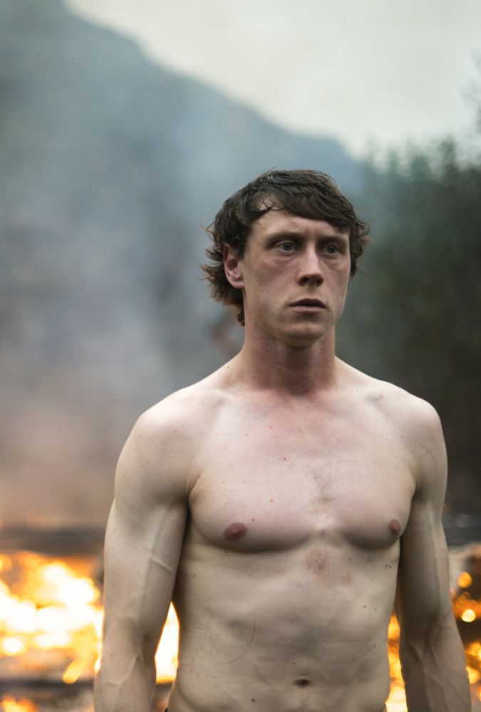 A shirtless man stands outdoors during daytime in front of a fire