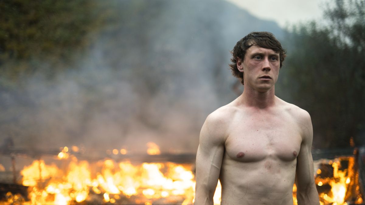 A shirtless man stands outdoors during daytime in front of a fire