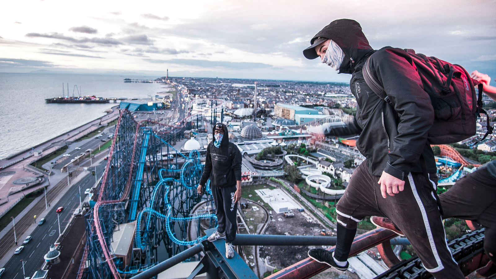 Two people with their faces covered wearing black tracksuits balance on skinny beams high in the sky - they appear to be on top of a tall building. Far below them are the tracks of rollercoasters in a theme park.