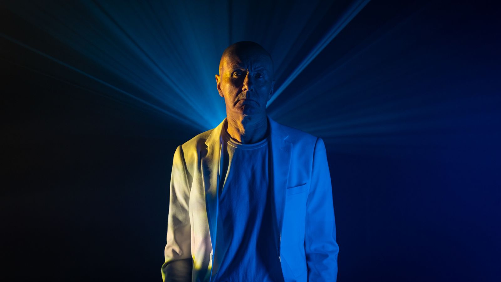 Irvine Welsh stands in a dark room wearring a white suit.