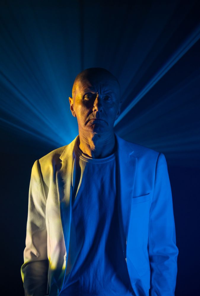 Irvine Welsh stands in a dark room wearring a white suit.
