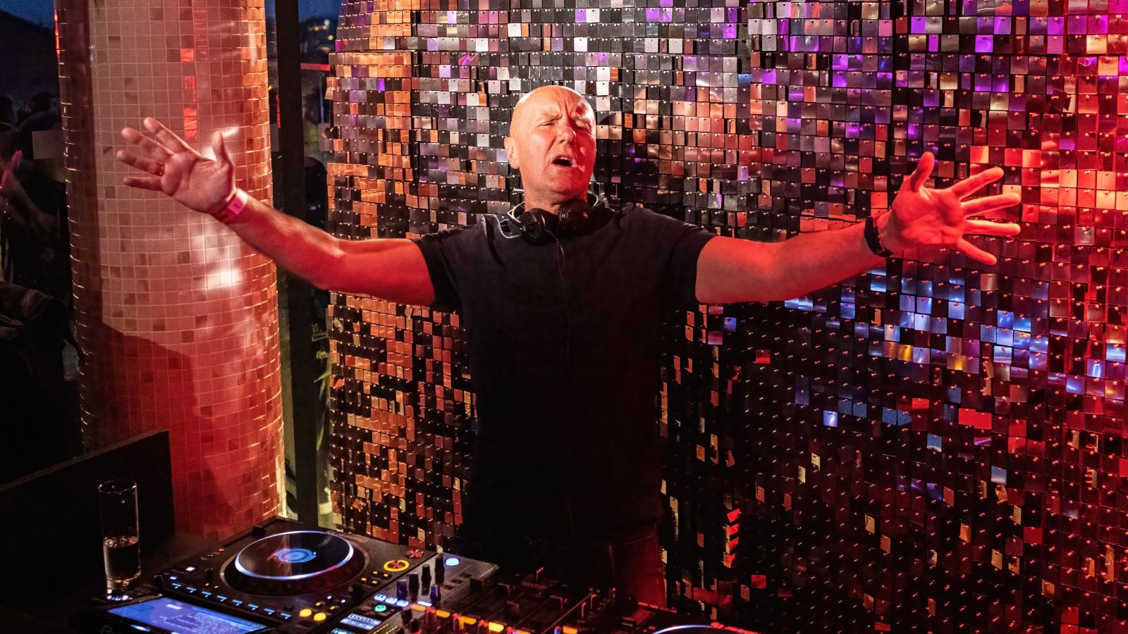 Irvine Welsh stands behind a dj deck underneath a disco ball.
