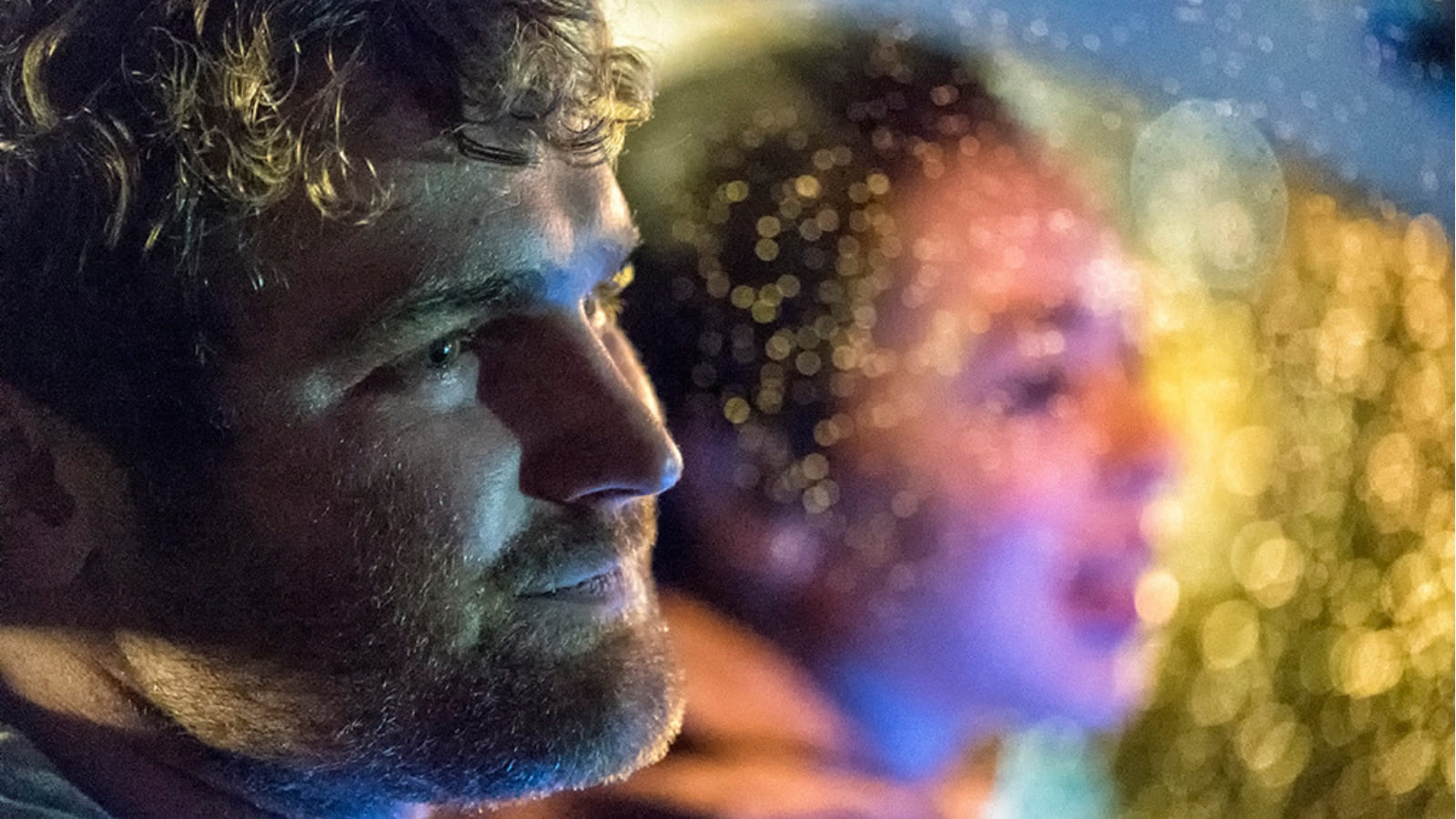 A man sits in a car alongside a woman, with yellow light shining behind them