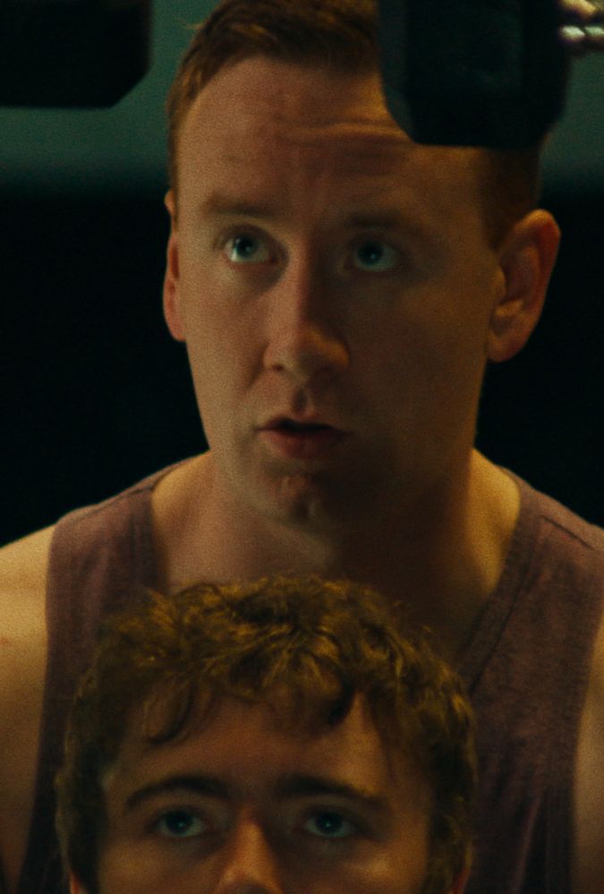 A man in a sleeveless shirt looks upward with a focused expression, while partially obscured by another person in front of him.