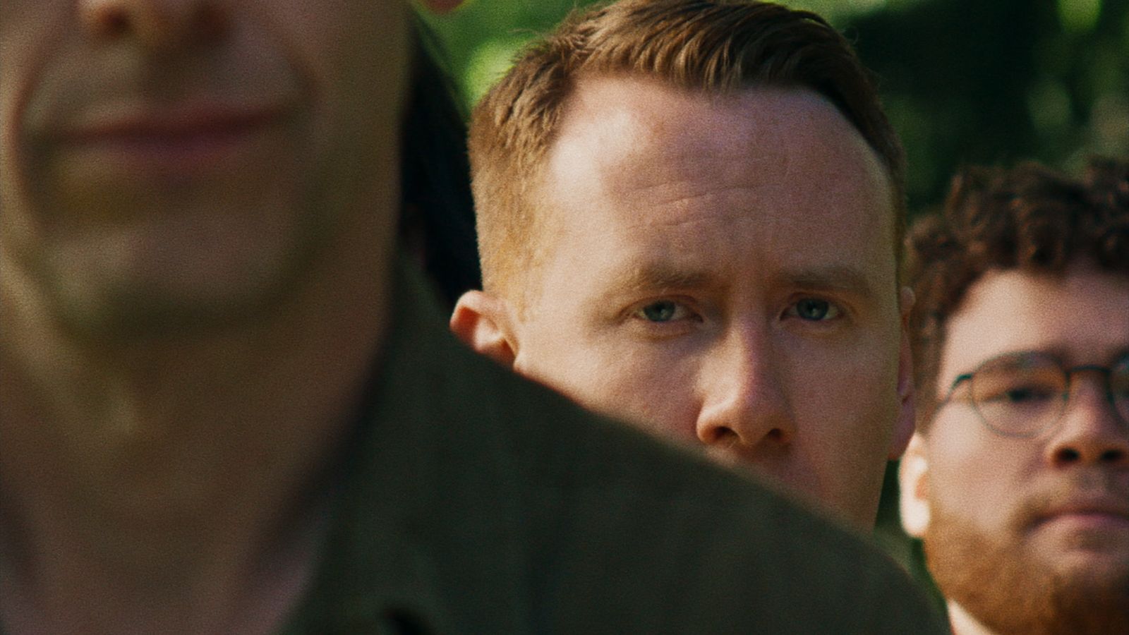Close-up of a man with short blond hair peering from behind another person, with intense focus. A second individual with curly hair and glasses appears in the background, slightly out of focus. The setting is outdoors with a blurred green backdrop.
