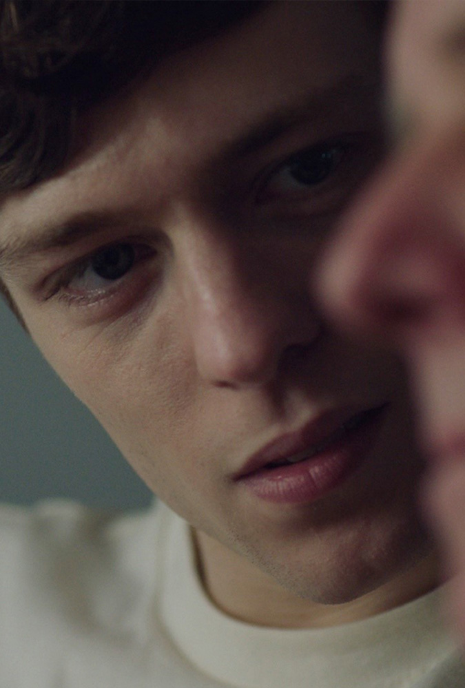 A close up shot of young man who sits in a white tshirt in a dim room, looking intently at at an older man who is closer to the camera - the focus is on the young man, who is Sebastian, a writer