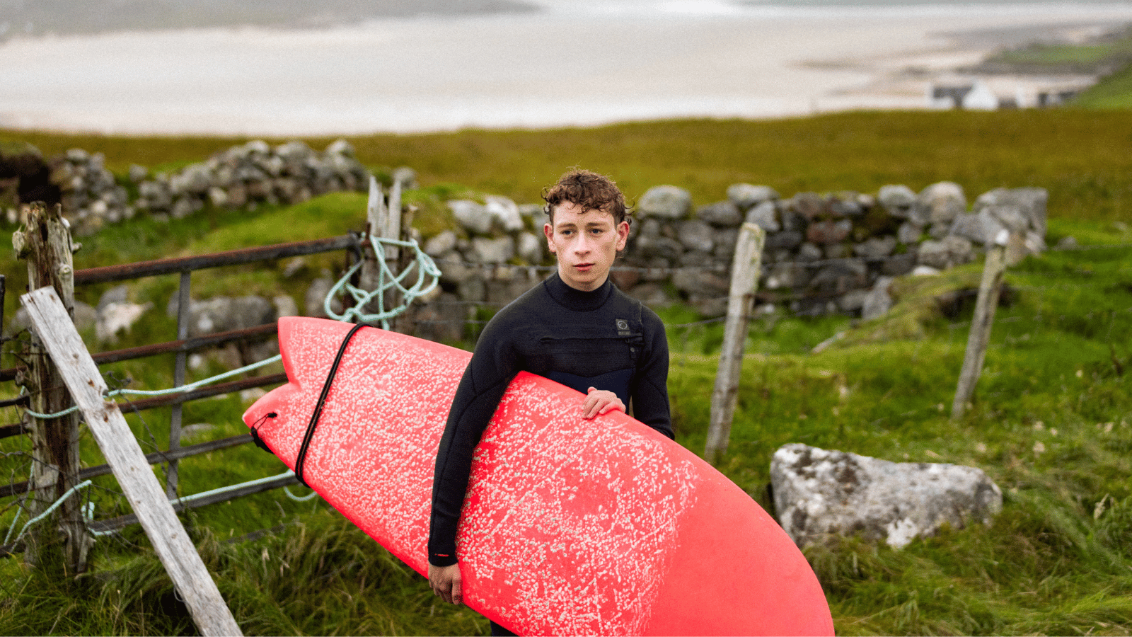 Still from silent Roar. Teenage boy in wetsuit holding red surf board.