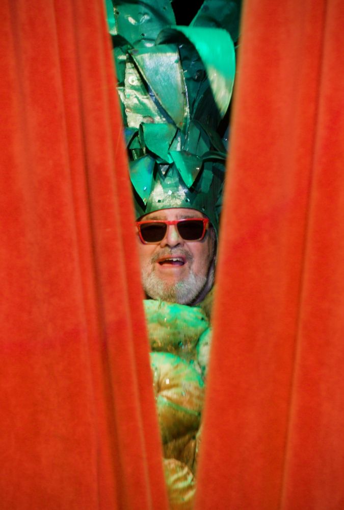 A man wearing a pineapple hat and sunglasses peaks through red stage curtains