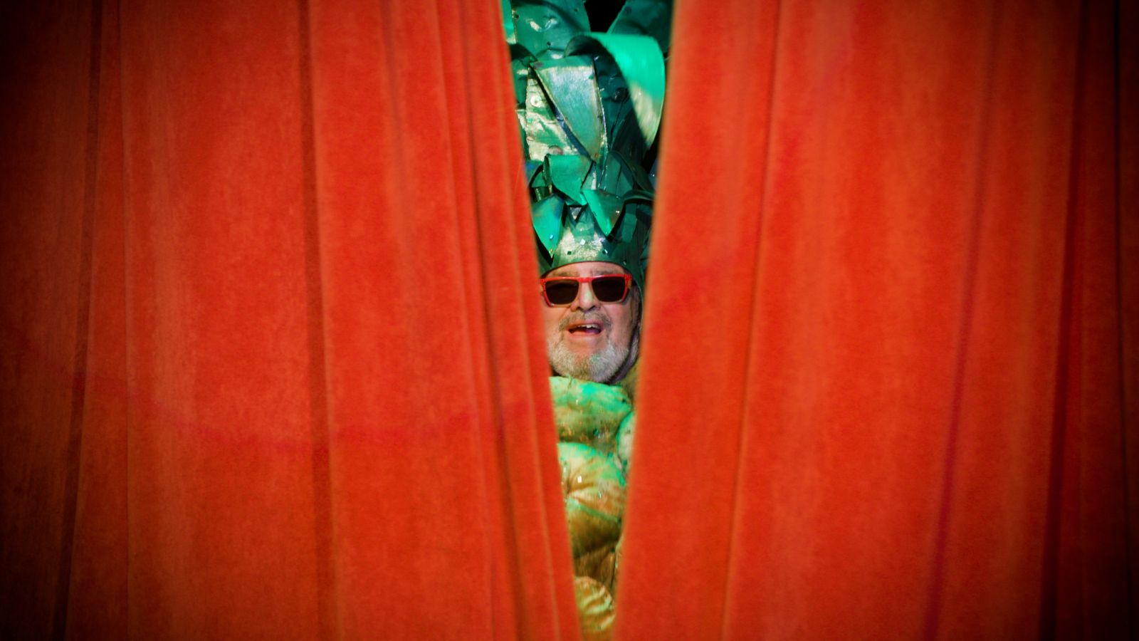 A man wearing a pineapple hat and sunglasses peaks through red stage curtains