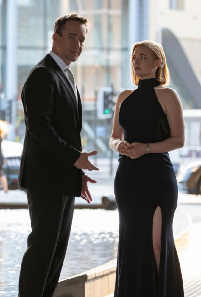 Succession Still where two characters stand in formal dress outside next to a water feature