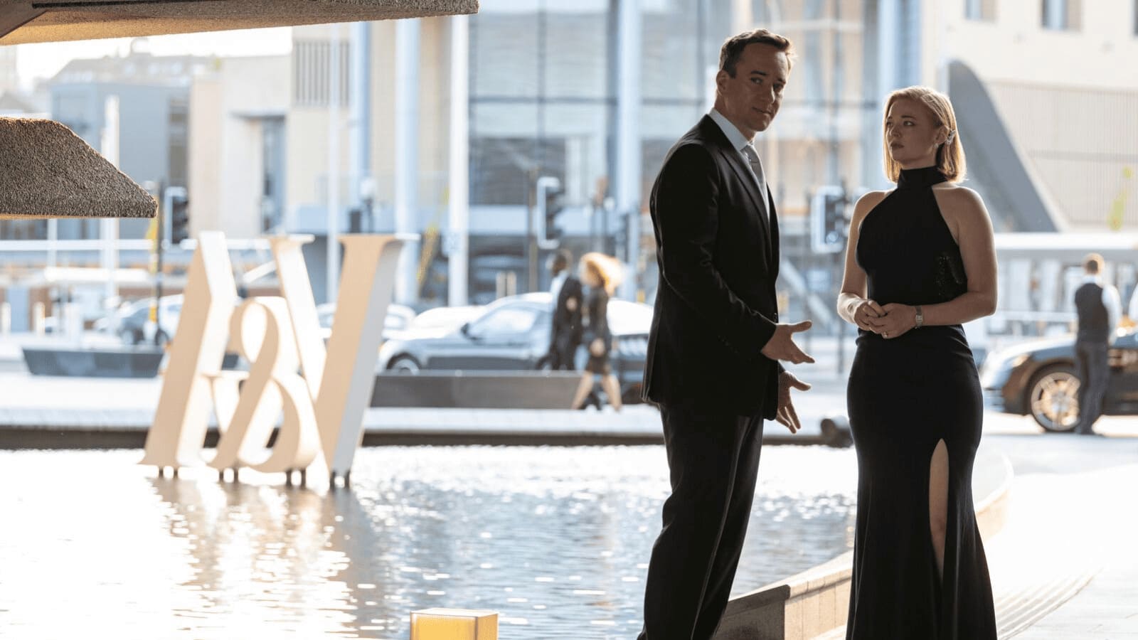 Succession Still where two characters stand in formal dress outside next to a water feature