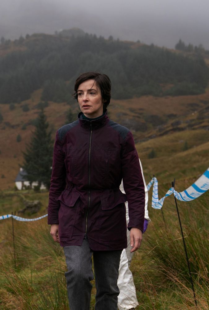 A woman in a dark jacket walks along a grassy hillside marked with blue and white striped tape. The background features misty, forested hills and a distant white house. The atmosphere is overcast and moody.
