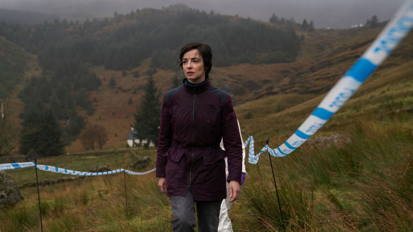 A woman in a dark jacket walks along a grassy hillside marked with blue and white striped tape. The background features misty, forested hills and a distant white house. The atmosphere is overcast and moody.
