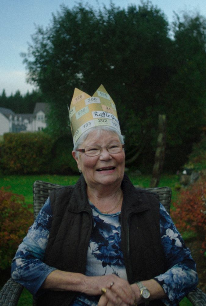 Elderly woman smiling outdoors, wearing a paper crown made of raffle tickets, sitting on a wicker chair against a garden backdrop.