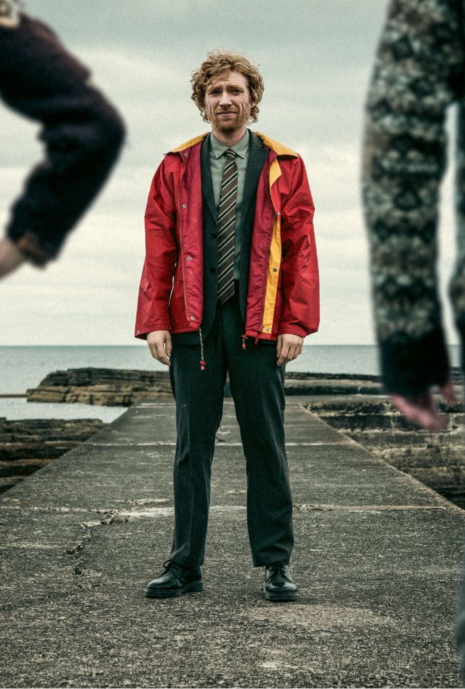 A man in a red jacket and tie stands on a weathered pier by the sea, framed by the blurred figures of two people in patterned sweaters. The sky is overcast, adding a moody atmosphere to the scene.