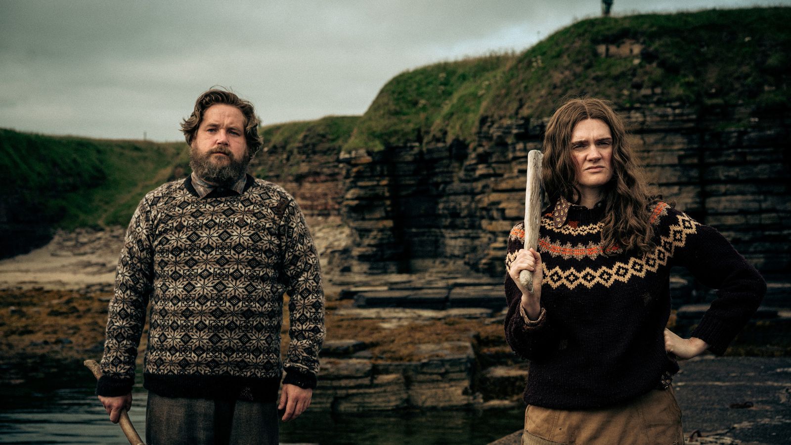 A man and a woman stand outdoors against a backdrop of rocky cliffs and overcast skies. Both wear intricately patterned wool sweaters. The man has a beard and holds a stick. The woman, with long hair, holds a piece of wood over her shoulder, striking a confident pose. landscape.