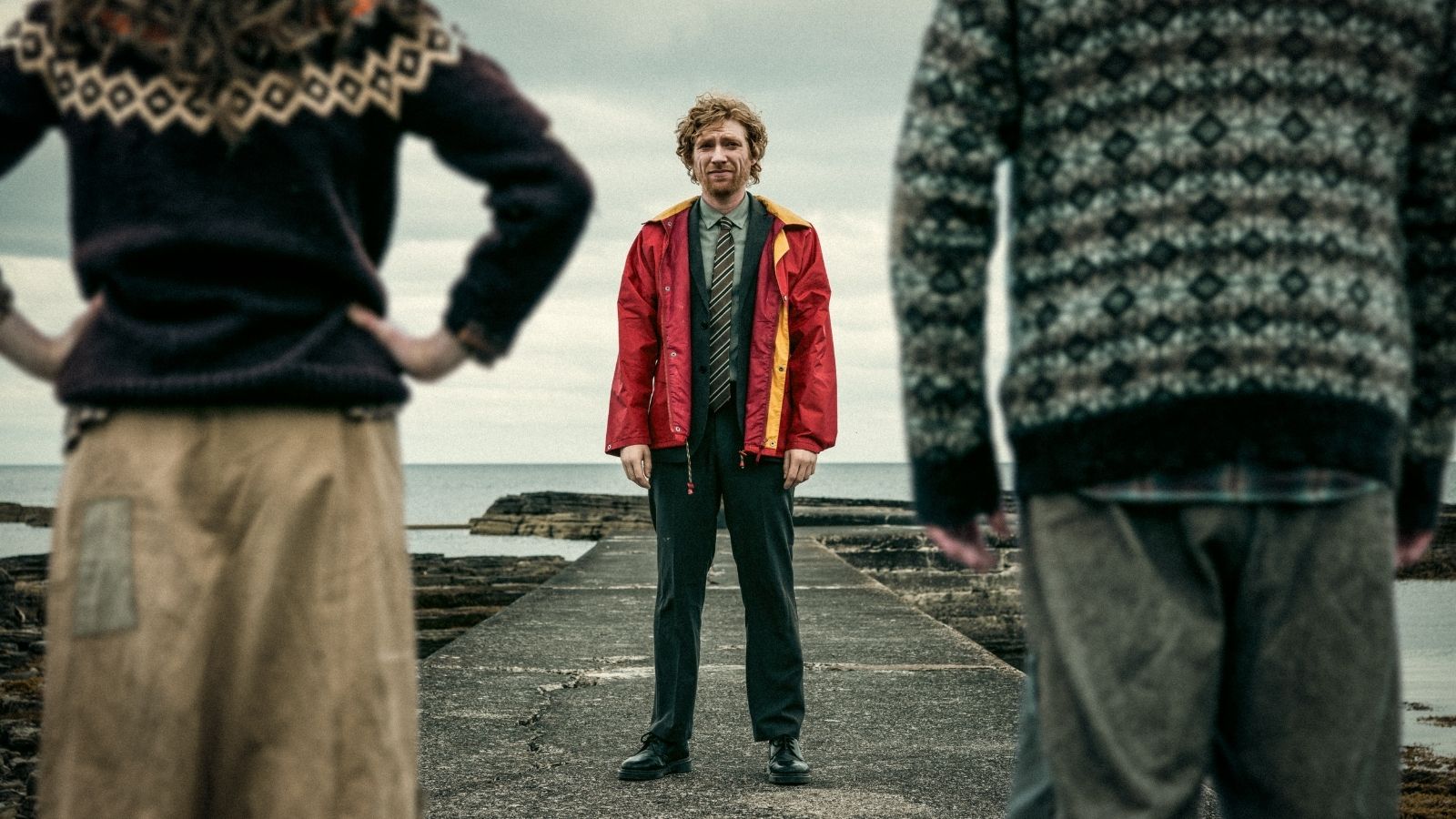A man in a red jacket and tie stands on a weathered pier by the sea, framed by the blurred figures of two people in patterned sweaters. The sky is overcast, adding a moody atmosphere to the scene.