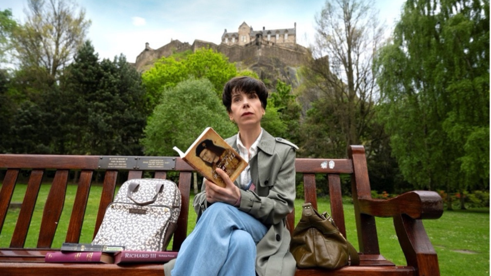 A woman with short dark brown hair in a light green coat and jeans (Actress Sally Hawkins) sits on a wooden bench reading a book about Richard III in a leafy park. Behind her is an old castle sitting a rocky hill - it is Edinburgh castle.