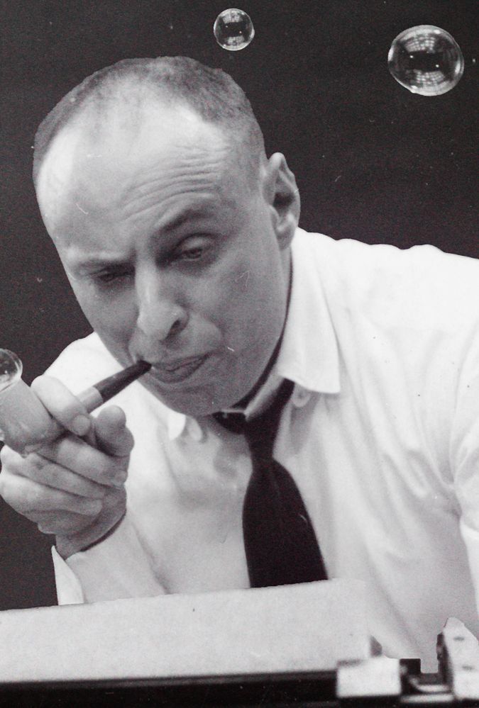 A man in a white shirt and black tie blows bubbles from a pipe while sitting at a typewriter. His expression is playful, surrounded by floating bubbles against a dark background.