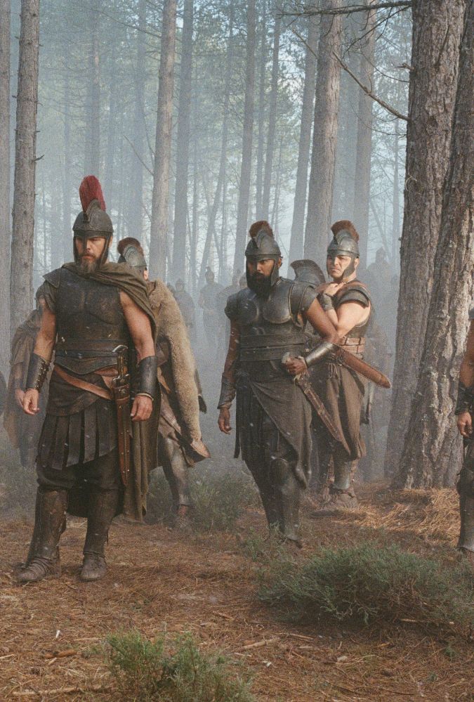 A group of warriors, dressed in ancient-style armour, walk through a hazy pine forest. The central figure, a bearded man, wears a helmet topped with a red crest, dark cuirass, a cape, and greaves. To his left and right, other soldiers march in matching gear, wearing helmets, breastplates, and carrying swords. The warriors have serious expressions. Tall, slender pine trees rise around them, with a light mist or smoke filling the background, casting a soft, diffuse light over the scene. The ground is covered in brown pine needles and sparse green shrubs.