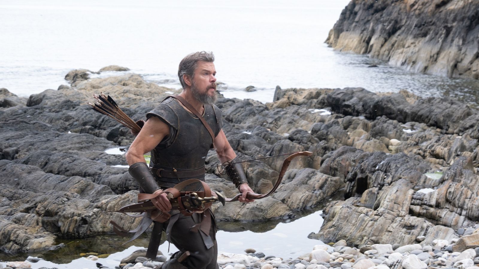 An archer with a beard and greying hair, wearing dark armour, bracers, and a quiver of arrows on his back, walks across a rugged, rocky shore. He holds a wooden bow in his left hand as he moves over the wet, dark stones near a calm body of water.