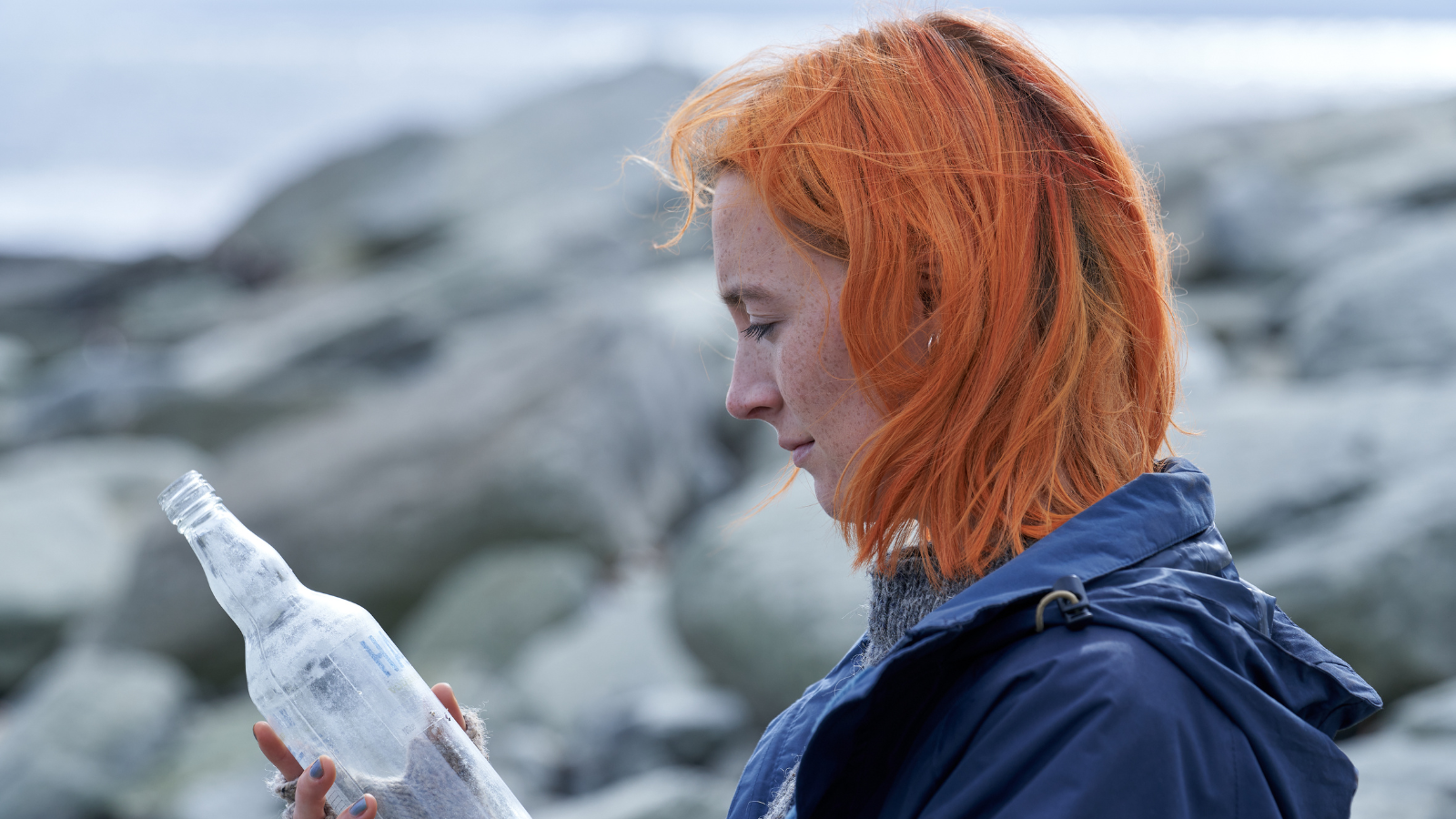 A young woman with bright orange hair examines a clear glass bottle, standing against a backdrop of blurred rocks. She wears a blue jacket and appear contemplative.