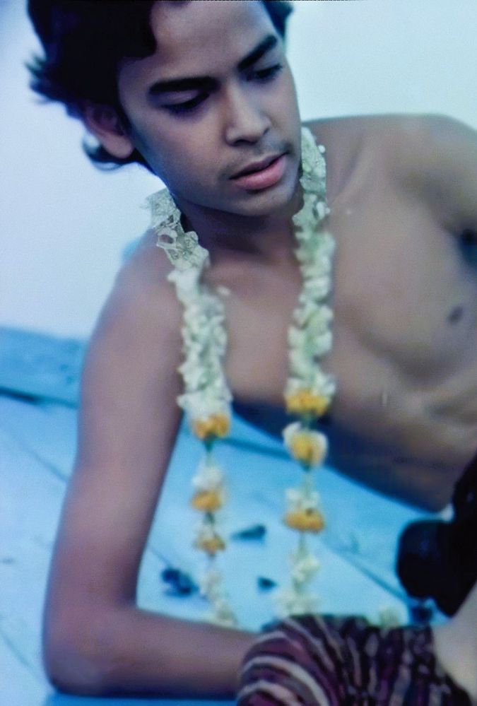 A young person with dark hair is leaning sideways, wearing a yellow and white flower garland around their neck. The background is a soft blue, giving the scene a serene feel. Their expression is calm and introspective.