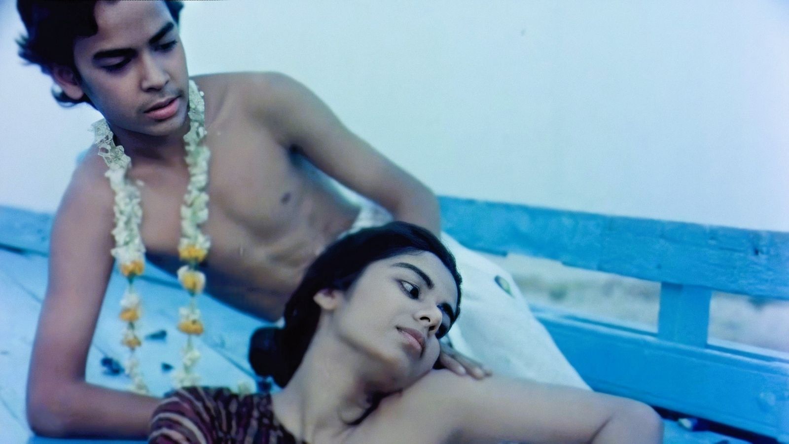 A shirtless young man wearing a garland of flowers reclines on a blue bench, gazing softly at a woman resting her head on his arm. The woman looks outward with a serene expression. The scene is bathed in a soft, cool light, creating a tranquil, intimate atmosphere.