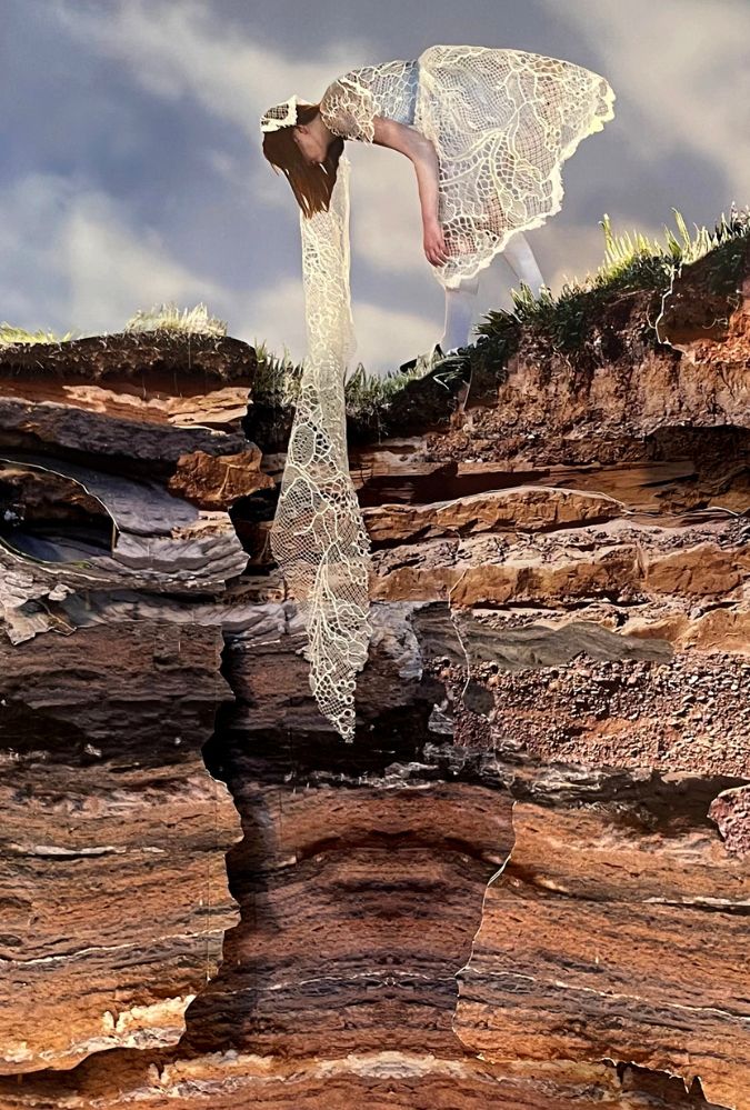 A person in a delicate, white lace dress leans over the edge of a rugged cliff, with fabric cascading down the layered, rocky surface. The backdrop features a blue sky with soft, wispy clouds.