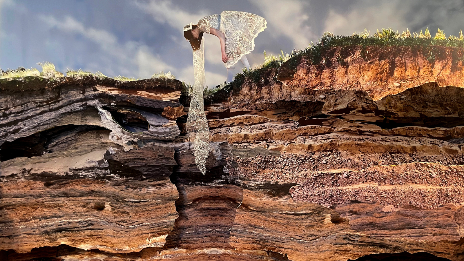 A person in a delicate, white lace dress leans over the edge of a rugged cliff, with fabric cascading down the layered, rocky surface. The backdrop features a blue sky with soft, wispy clouds.