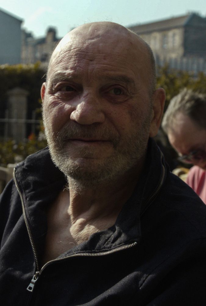 An outdoor close-up shot of an older bald man with a short grey beard, wearing a black zip-up jacket.