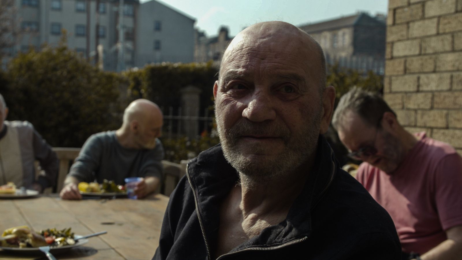 An outdoor close-up shot of an older bald man with a short grey beard, wearing a black zip-up jacket. In the blurred background, other men sit around a wooden table in a sunny garden setting.