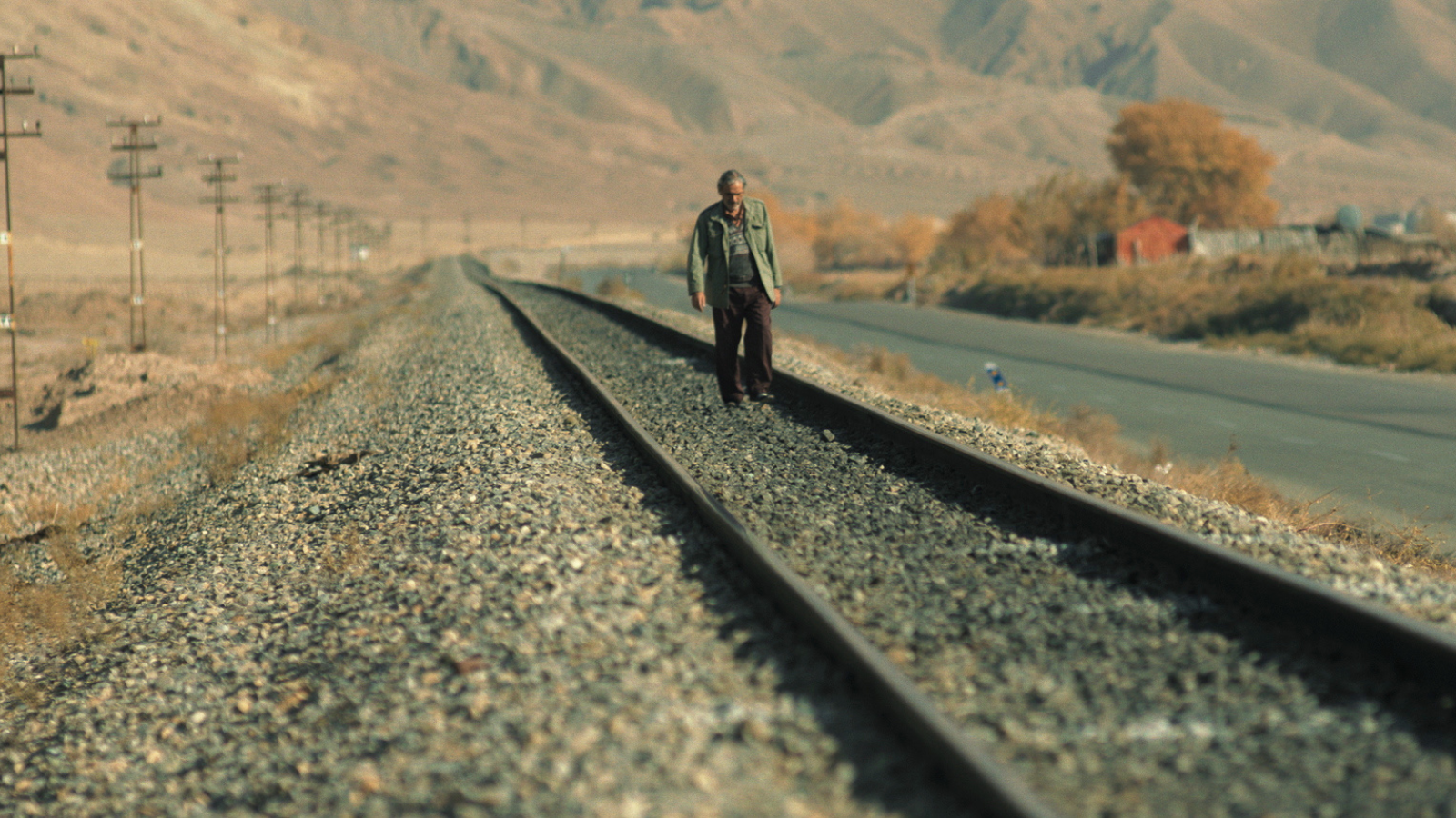 An old man wanders empty railroad tracks along a dusty highway