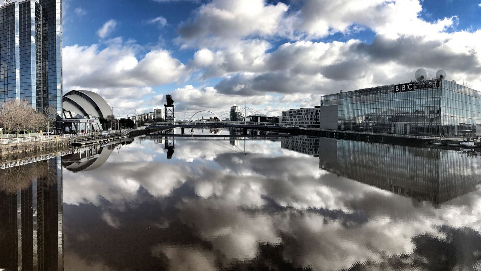A wide panoramic shot of the River Clyde in Glasgow on a bright day. The calm, mirror-like water perfectly reflects a dramatic blue sky filled with puffy, white cumulus clouds. On the left bank stands the glass facade of the Crowne Plaza hotel, next to the curved, silver dome of the SEC Armadillo. Further down the river, the Millennium Bridge spans the water, with the tall Finnieston Crane visible in the distance. On the right bank, the large, glass BBC Scotland building sits prominently at the water's edge. The entire cityscape and sky are symmetrical, mirrored upside down in the still river below.