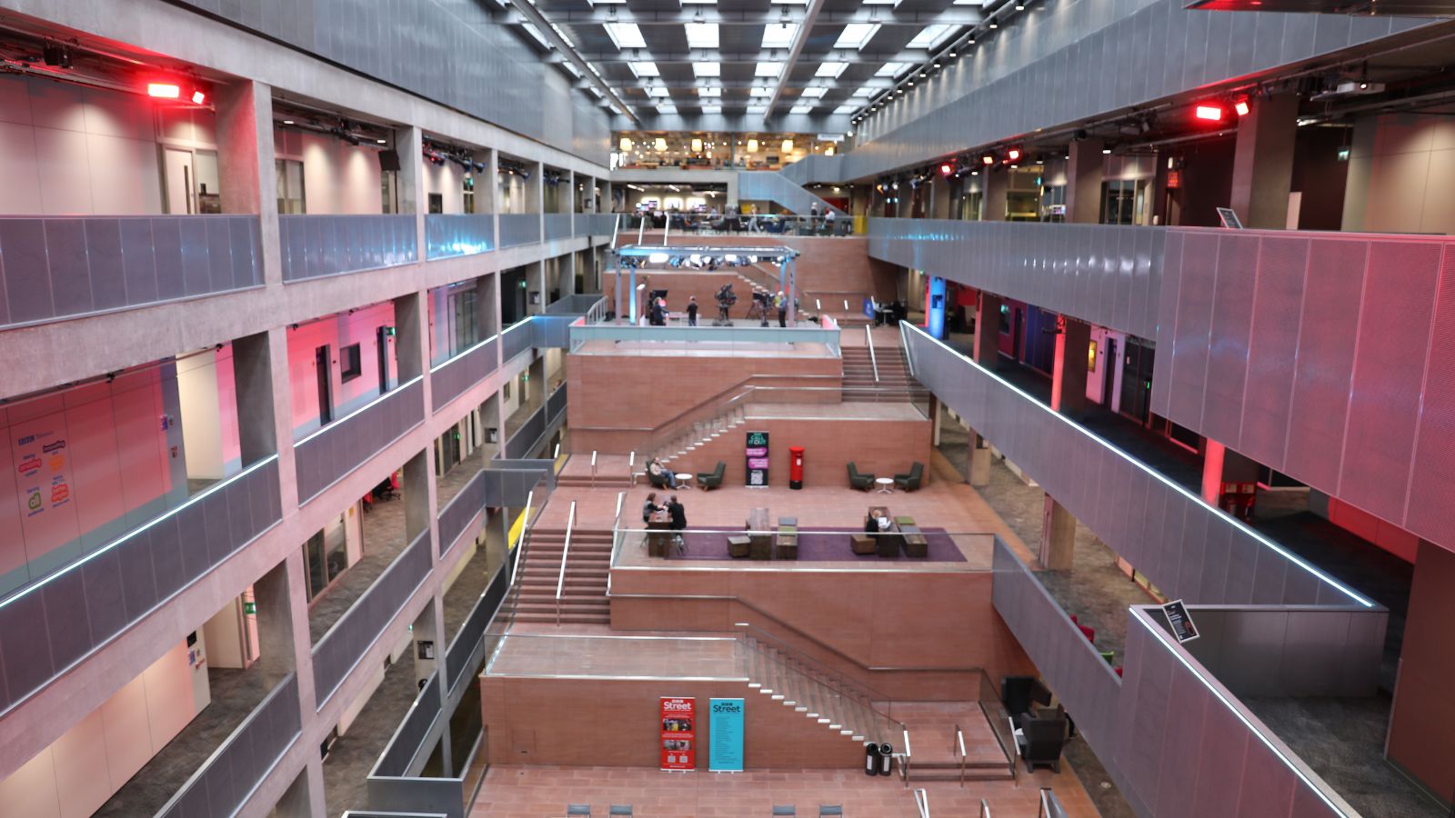 An elevated, wide-angle shot of a multi-story, modern atrium. Multi-level concrete balconies line both sides of the atrium under a skylight grid. Stairs on a central structure made of reddish-brown brick transition from level to level. Red accent lighting illuminates various parts of the space. People are scattered on different levels of the central staircase structure, some sitting in lounge areas on the terraces.