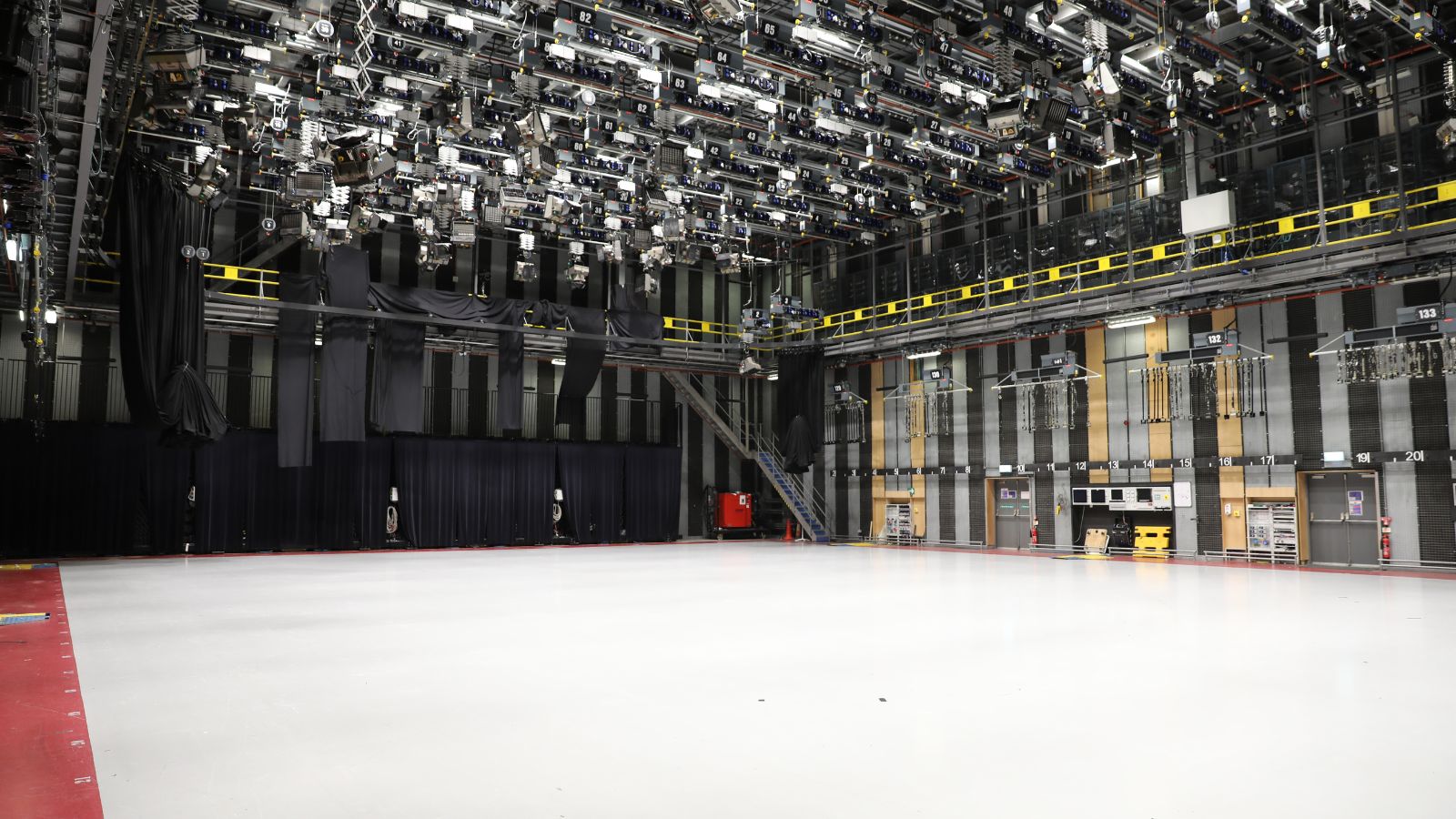 An expansive, empty television studio with a polished white floor and a complex overhead grid of stage lighting fixtures. Black acoustic curtains drape the left and back walls, while the right wall features vertical wooden panels, numbered positions, and control equipment. A metal staircase and gantry walkway run along the upper level.