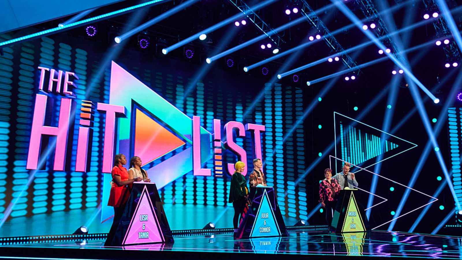 A wide shot of a brightly lit game show stage with three pairs of contestants standing behind podiums. The set is decorated with a vibrant, neon-coloured aesthetic, featuring the logo of "The Hit List" in large, stylized letters in the background. The logo features a play button icon. The stage is illuminated by multiple blue and purple spotlight beams crisscrossing the air. Each pair of contestants stands behind a triangular podium with their names printed on it.
