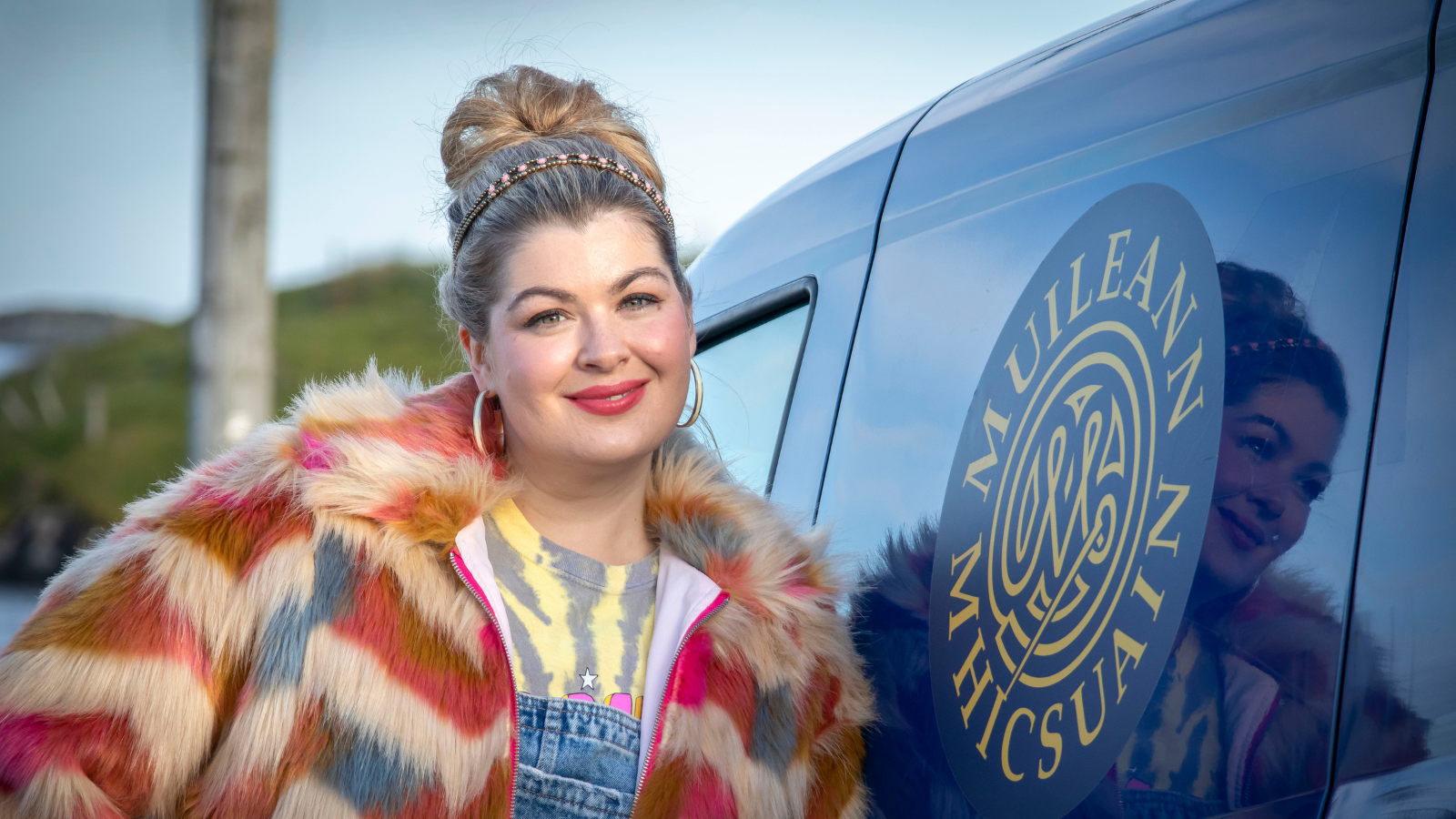 Still from An Clò Mòr. Woman with blonde hair and fluffy coat stands by a van outside.